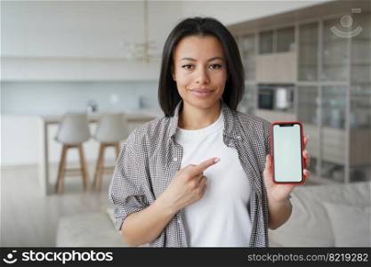 Female showing smartphone with mock up blank screen looking at camera, standing at home. Woman holding mobile phone, advertising new apps, recommending online store offer with discounts.. Female advertises mobile apps showing phone with mockup blank screen at home. Online store offer