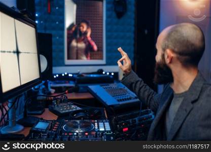 Female singer in recording room and sound engineer and at remote control panel in studio. Musician in headphones listens composition, professional music mixing