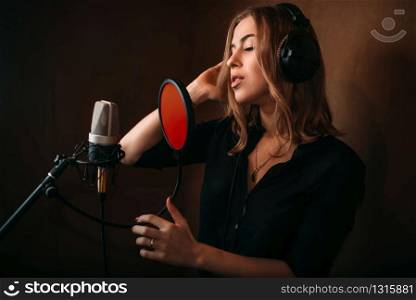 Female singer recording a song in music studio. Woman vocalist in headphones against microphone. Audio recording. Professional digital sound technologies. Female singer recording a song in music studio