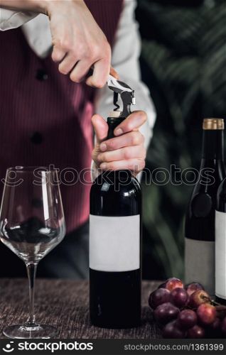 Female sommelier opening wine bottle with corkscrew. Fresh grapes and empty wine glass on table.