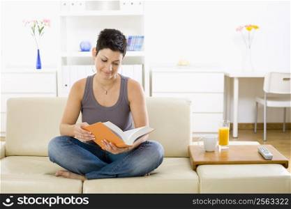 Female student sitting on sofa at home, reading book.