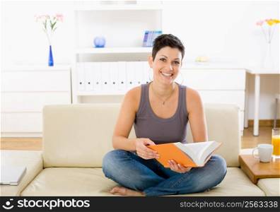 Female student sitting on sofa at home, reading book.