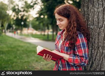 Female student studying the book in summer park. Ginger teenager leisures outdoors, pretty girl relax outside