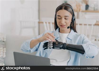 Female student with disability wearing headset learning at laptop at home. Disabled girl setting, touching her modern bionic prosthetic arm before work or distant education on computer.. Girl student with disability setting bionic prosthetic arm before work or learning at laptop at home