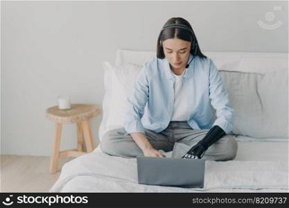 Female student with disability wearing headset studying online at laptop, typing, using bionic prosthetic arm, sitting on bed at home. Elearning, distant job of disabled people concept.. Female student with disability studying online at laptop, using bionic prosthetic arm sitting on bed