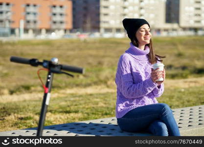 Female student with electric scooter taking a coffee break enjoying the winter sunlight with her eyes closed. Lifestyle concept.. Female student with electric scooter enjoying the winter sunlight with her eyes closed.