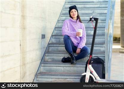 Female student with electric scooter taking a coffee break sitting on some steps outdoors. Lifestyle concept.. Female student with electric scooter taking a coffee break sitting on some steps outdoors