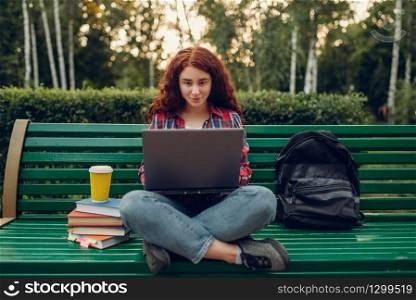 Female student with laptop sitting on the bench in yoga pose, summer park, front view. Ginger teenager leisures outdoors, pretty girl relax outside