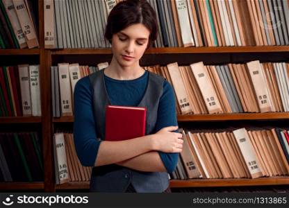 Female student with the book in hands standing with eyes closed in college library. Bookshelf with books and textbooks on the background.