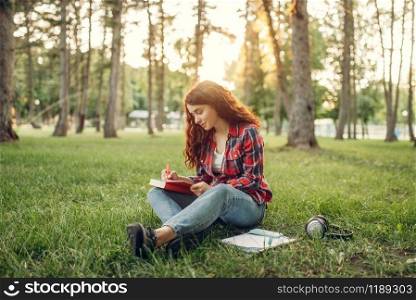Female student writing in notebook on the grass in summer park. Ginger teenager studying and leisures outdoors