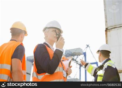 Female supervisor using walkie-talkie while workers discussing in background at shipyard