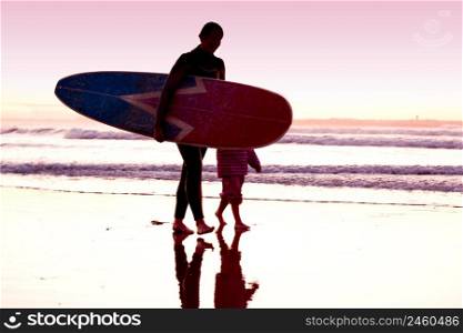 Female surfer and her daughter walking in the beach at the sunset