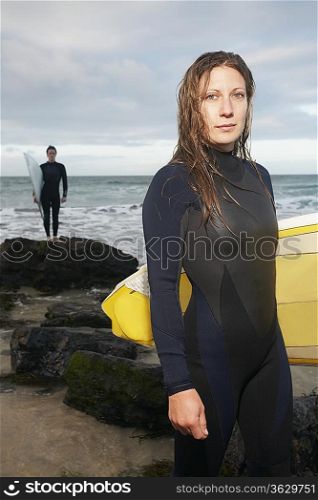 Female surfer carrying surfboard standing on beach, another surfer standing in background, portrait