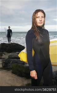 Female surfer carrying surfboard standing on beach, another surfer standing in background, portrait