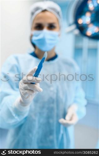 Female surgeon in gown and mask shows scalpel in operating room, lamp on background, surgery operation. Doctor in uniform, medical clinic worker, surgical medicine and health, healthcare in hospital. Female surgeon in gown and mask shows scalpel