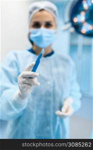 Female surgeon in gown and mask shows scalpel in operating room, lamp on background, surgery operation. Doctor in uniform, medical clinic worker, surgical medicine and health, healthcare in hospital. Female surgeon in gown and mask shows scalpel