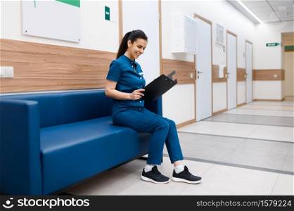 Female surgeon with notebook sitting on sofa in clinic hall, surgery. Doctor in uniform, medical worker, medicine and health, professional healthcare in hospital. Female surgeon sitting on sofa in clinic hall