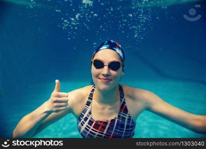 Female swimmer in swimsuit, swimming cap and glasses shows thumbs up underwater in pool. The woman holds her breath in the water, breath-hold technique exercise. Female swimmer shows thumbs up underwater in pool