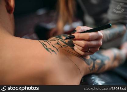 Female tattooist drawing tattoo on male shoulder. Professional tattooing in salon