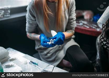 Female tattooist in blue sterile gloves prepares tattoo machine, master in salon. Professional tattooing in studio. Female tattooist prepares tattoo machine