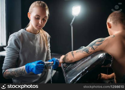 Female tattooist makes tattoo by machine on the shoulder. Professional tattooing in salon. Tattooist makes tattoo by machine on male shoulder