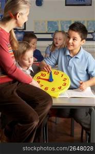 Female Teacher In Primary School Teaching Children To Tell Time In Classroom