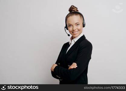 Female technical support worker in dark suit with black wireless headset standing sideways with her arms crossed and helping caller to resolve issues, woman in headphones posing on light background. Friendly business woman in headset in dark suit ready for web conference