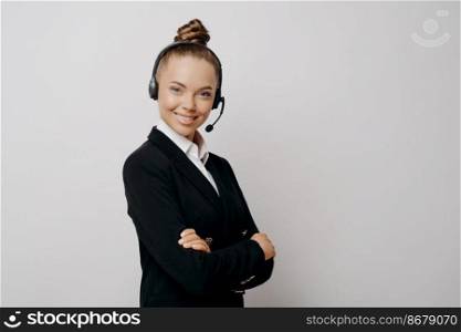 Female technical support worker in dark suit with black wireless headset standing sideways with her arms crossed and helping caller to resolve issues, woman in headphones posing on light background. Friendly business woman in headset in dark suit ready for web conference