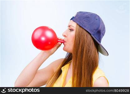 Female teenager inflating red balloon.. Time for party. Young trendy woman prepare birthday celebration decoration. Teenage girl in cap blowing red balloon.