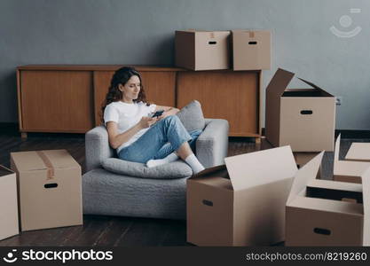 Female tenant or homeowner preparing for relocation orders moving service using smartphone sitting in armchair surrounded by cardboard boxes. Advertisement of removal, delivery company.. Female orders moving service using phone app on relocation day sitting in room with carton boxes