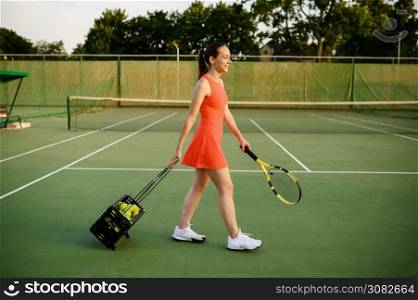 Female tennis player carries basket with balls on outdoor court. Active healthy lifestyle, sport game competition, fitness training with racquet. Female tennis player carries basket with balls