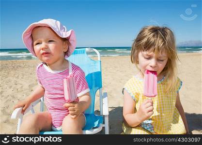 Female toddler and sister eating ice lollies on beach