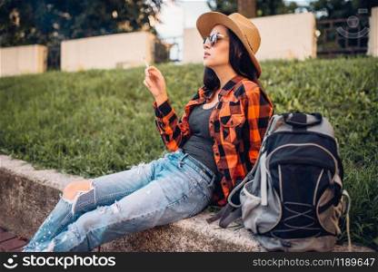 Female tourist in hat and sunglasses smoke cigarettes, break during the tour. Summer adventure of young woman, city walking. Female tourist in sunglasses smoke cigarettes