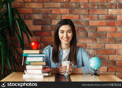 Female university student sitting at the table with textbooks and apple on the top, brick wall on background, knowledge concept. Young woman with books