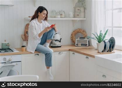Female uses modern phone, messaging online at kitchen at home, rests after chores. Woman housewife holding smartphone, shopping on internet, ordering groceries online, using delivery service app.. Female shopping groceries online by mobile phone app, sitting in modern kitchen at home. E commerce