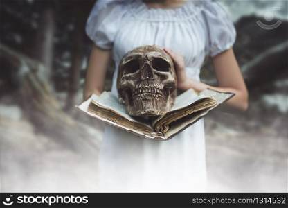 Female victim in white dress holds book and human skull in hand, forest on background. Photo in horror style, exorcism. Female victim holds book and human skull in hand