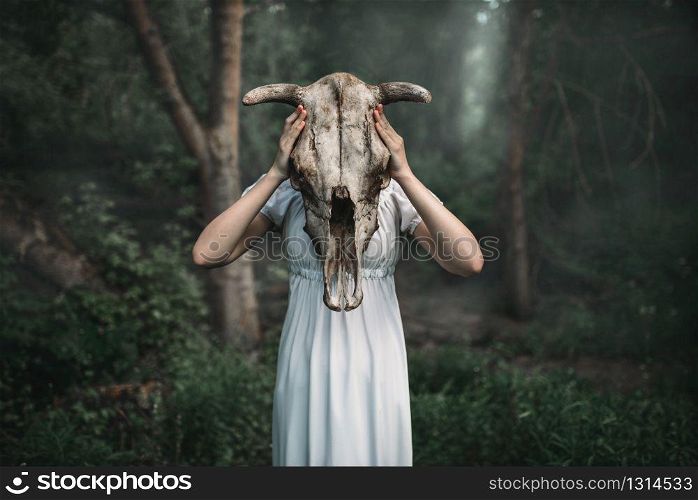 Female victim in white dress with skull of the animal instead of the head, forest on background. Photo in horror style, exorcism. Victim with skull of the animal instead of head
