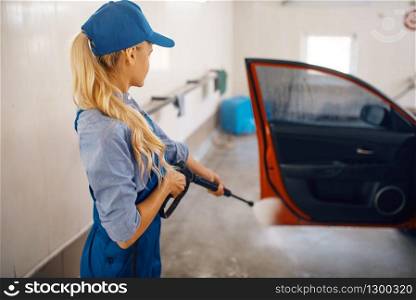 Female washer in uniform cleans door with high pressure gun in hands, car wash. Woman washes vehicle, carwash station, car-wash business. Female washer cleans door with high pressure gun