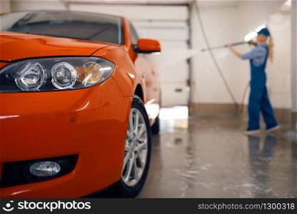 Female washer in uniform cleans wheel with high pressure gun in hands, car wash. Woman washes vehicle, carwash station, car-wash business. Female washer cleans wheel with high pressure gun