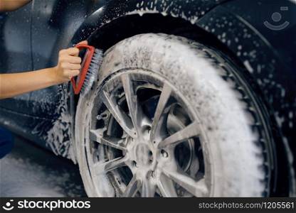 Female washer with brush in hand cleans wheel in foam, car wash. Woman washes vehicle, carwash station, car wash business. Female washer with brush cleans wheel, car wash