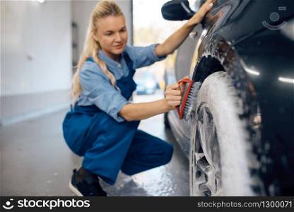Female washer with brush in hand cleans wheel in foam, car wash. Woman washes vehicle, carwash station, car-wash business