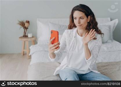 Female waving hand at phone camera, greeting, talking on videocall, sitting in bedroom. Hispanic woman answering call, welcoming interlocutor during video chat at home. Online communication concept.. Female waving hand at phone camera, greeting, talking on video call in bedroom. Online communication