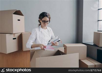 Female wearing glasses holding notebooks packing cardboard boxes on moving day, focused hispanic businesswoman employee packs things in carton, preparing for relocation in new office.. Businesswoman packs things in cardboard boxes on moving day, preparing for relocation in new office