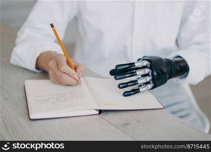 Female with disability makes notes, writing with pencil, holding notebook using robotic hand, close-up. Disabled woman writes in diary, sitting at table. Advertising of bionic prosthesis.. Female makes notes writes in diary, holds notebook using robotic hand. Bionic prosthesis advertising