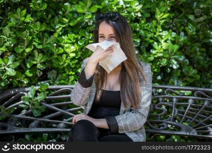 Female with pollen allergy blowing nose in paper tissue while sitting on bench near lush bush in park. Woman with hay fever blowing nose in park