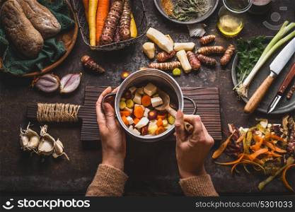 Female woman hands holding pan with diced colorful vegetables and a spoon on rustic kitchen table with vegetarian cooking ingredients and tools. Healthy and clean food cooking and eating concept.