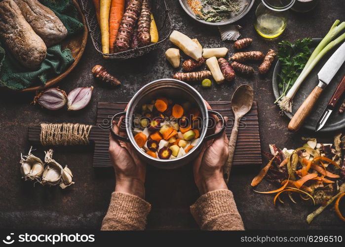 Female woman hands holding pan with diced colorful vegetables on dark rustic kitchen table with vegetarian cooking ingredients and tools. Healthy and clean seasonal food cooking and eating concept