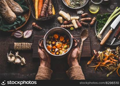 Female woman hands holding pan with diced colorful vegetables on dark rustic kitchen table with vegetarian cooking ingredients and tools. Healthy and clean seasonal food cooking and eating concept