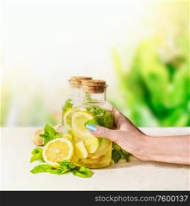 Female women hand holding bottle with ginger, lemon, mint lemonade on white table at green nature background. Healthy summer drinks. Natural Immun booster. Infused fruits water.