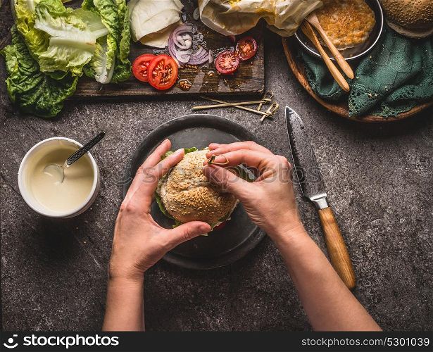 Female women hands holding homemade tasty burger on rustic kitchen table background with ingredients, top view